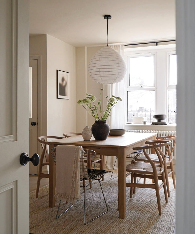 Light-filled dining room with a wooden table, mixed dining chairs, a large paper pendant light, and ceramic vases beside tall windows.
