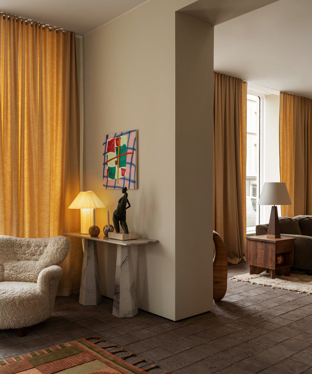 An open plan living area focusing a cosy corner featuring a table lamp on a console table with other decorative objects and a lounge chair in front upholstered in sheepskin