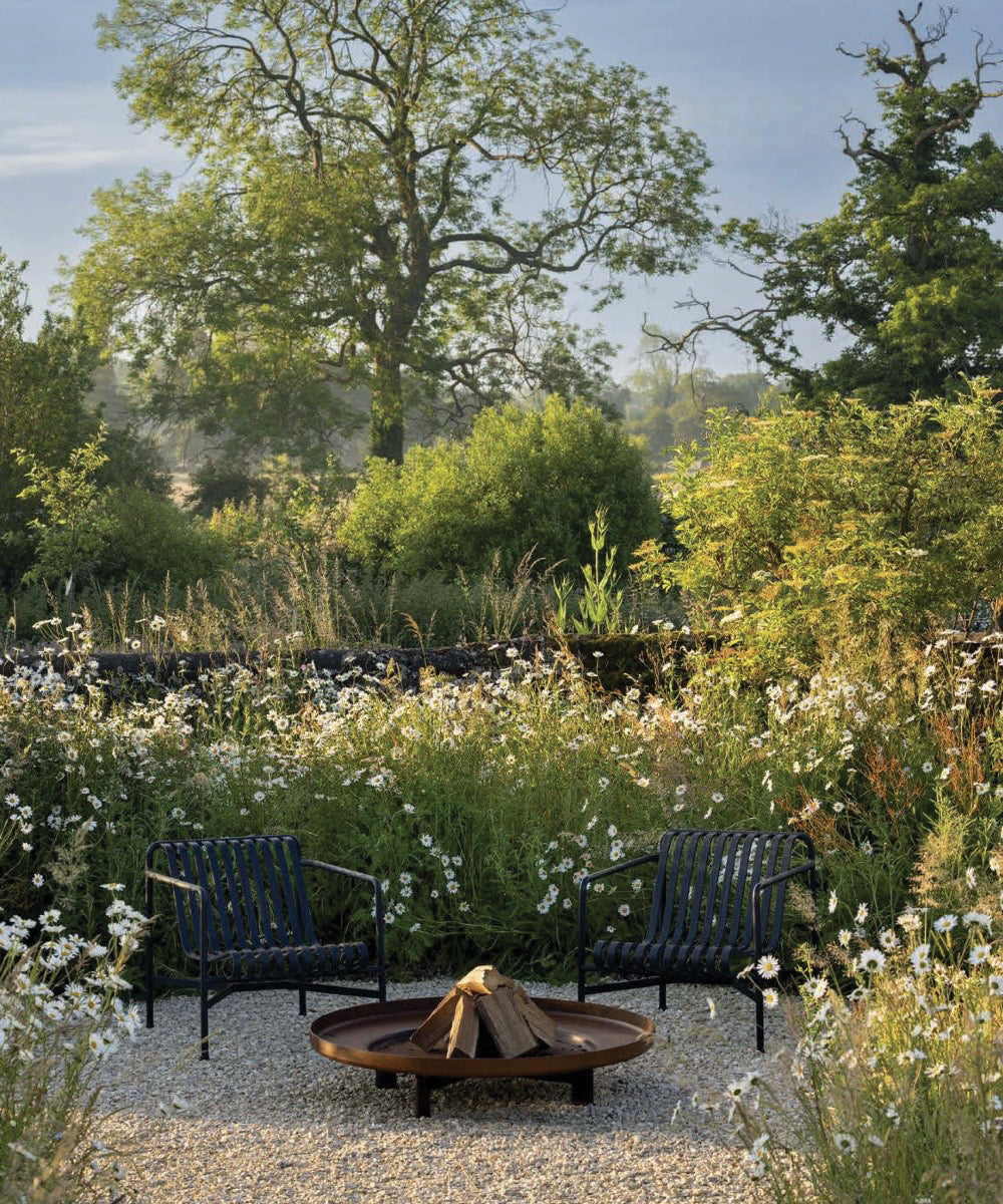 Two black chairs around a fire pit in a gravel seating area, surrounded by wildflowers and lush garden greenery.