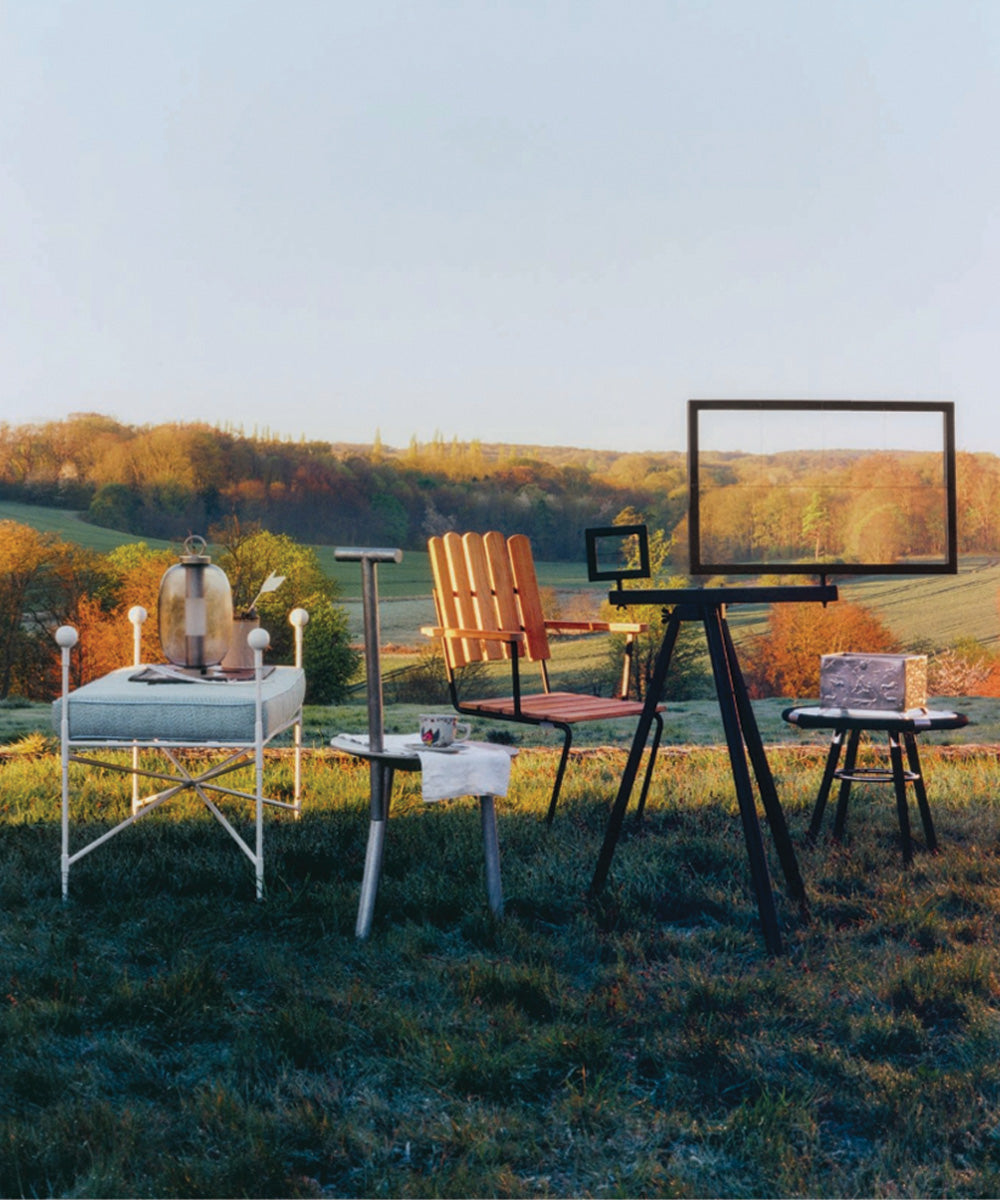 Curated selection of contemporary outdoor furniture arranged in a grassy countryside setting at sunrise, featuring chairs, tables, and lighting against rolling hills.