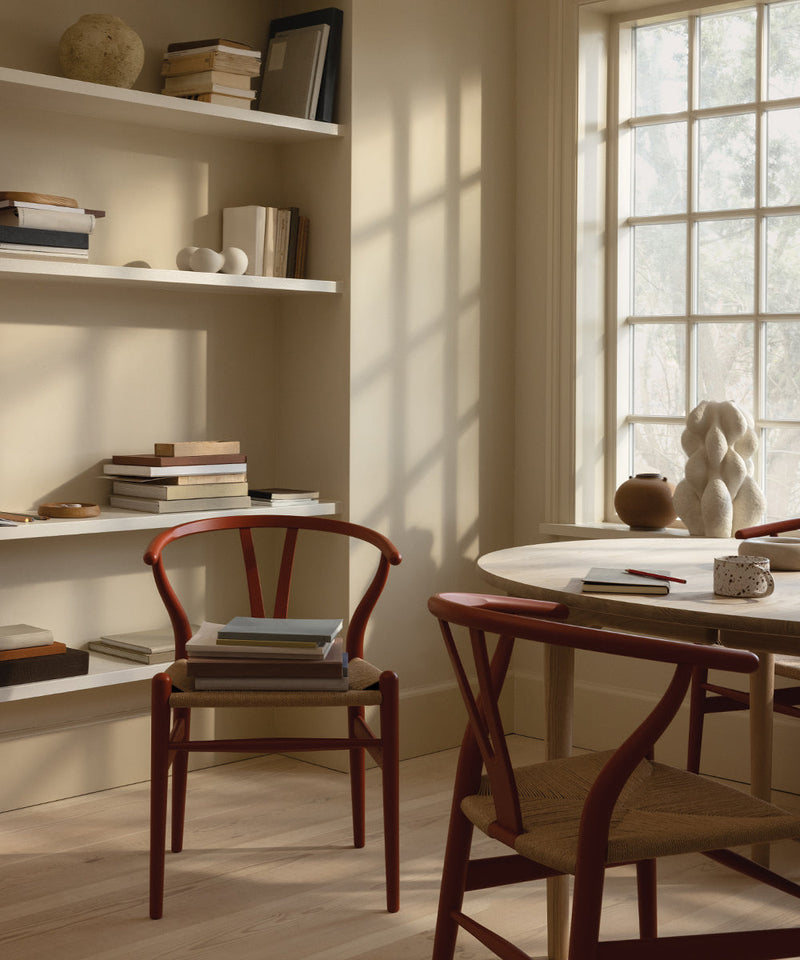 Sunlit dining nook with red wishbone chairs, a round wooden table, built-in bookshelves, and soft neutral decor.
